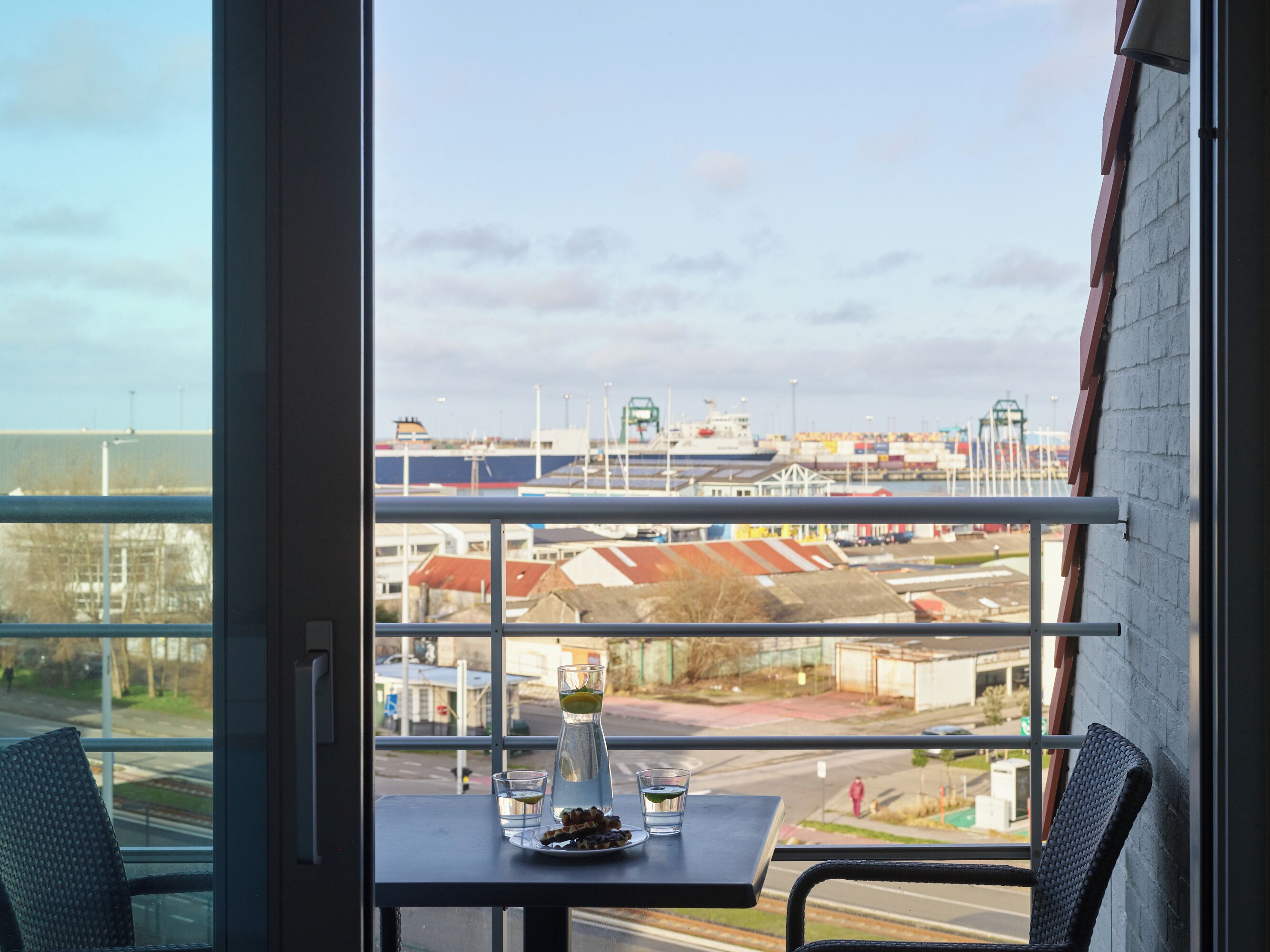 Une table avec un verre d’eau et une assiette de nourriture, une chaise, et une vue sur un port avec des navires et des bâtiments.