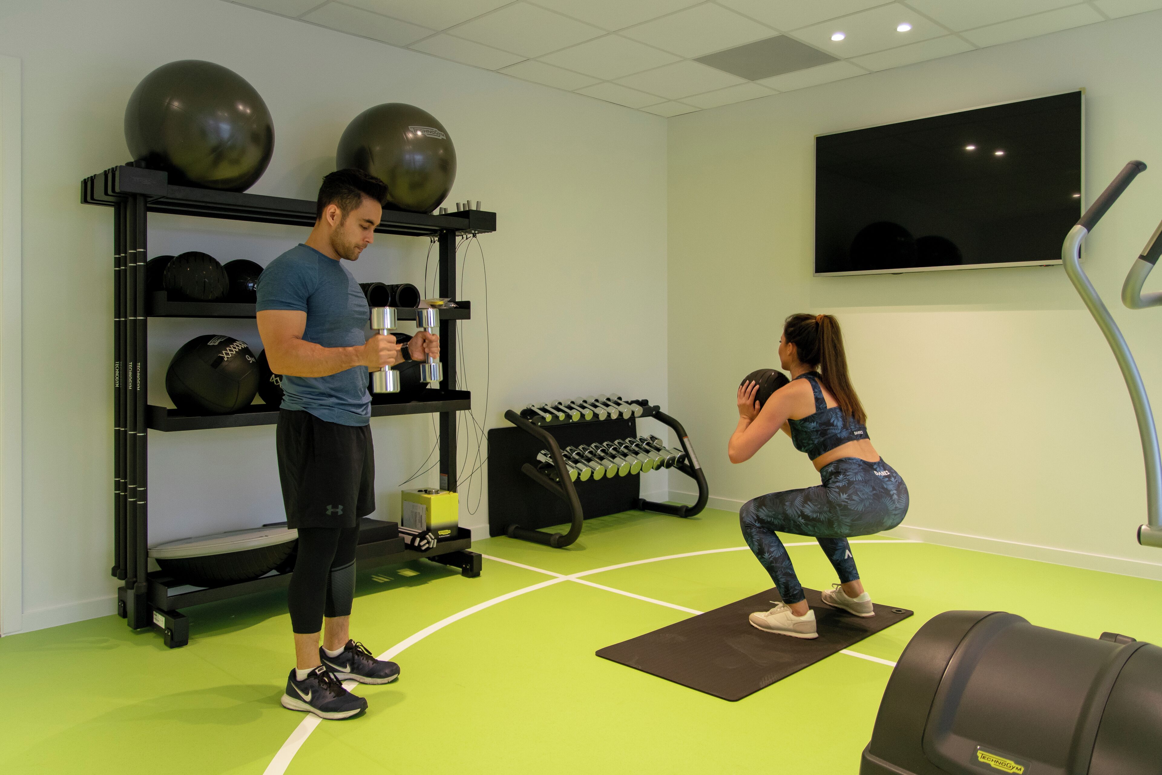 A gym with exercise equipment, a wall-mounted TV, and a green floor.
