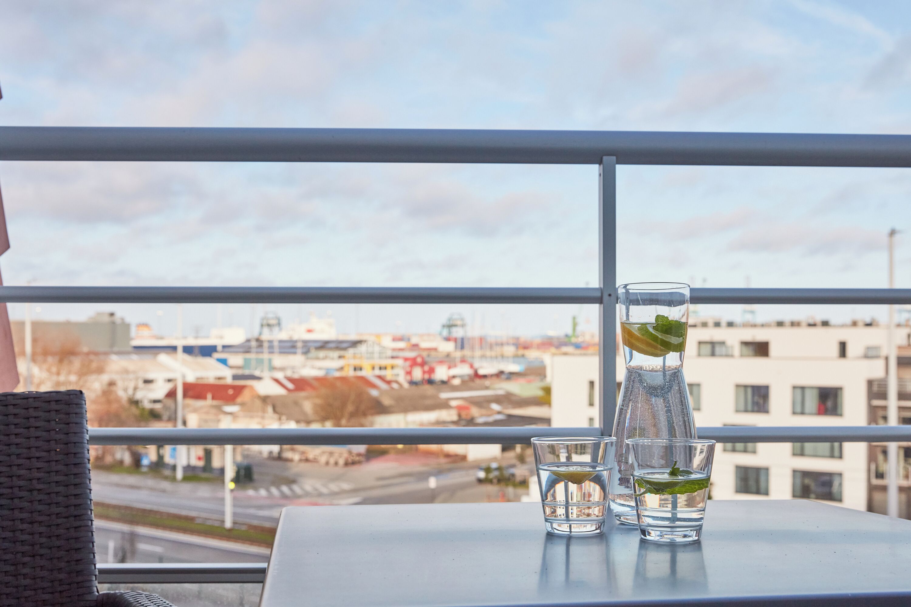 Ein Tisch mit einem Glas Wasser, einem Glas mit Limettenscheibe und einer Wasserflasche auf einem Balkon mit Blick auf eine Stadtlandschaft.