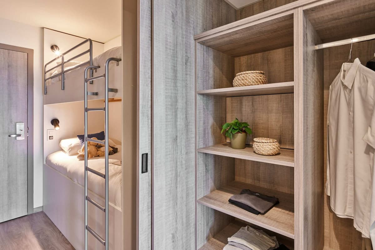 A modern bedroom with a bunk bed, a wardrobe with open shelves, and a bedside table with a lamp.