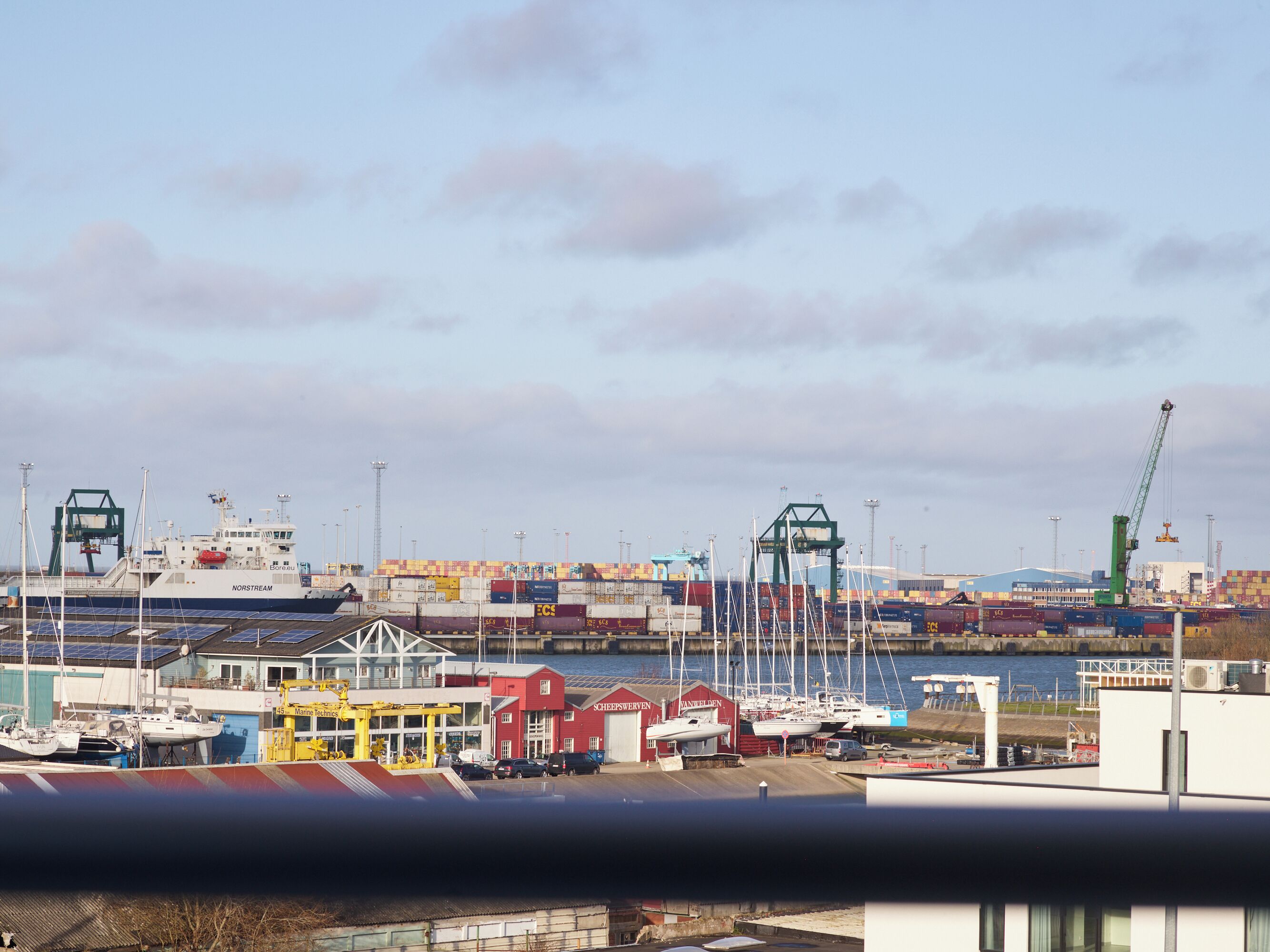 A harbor with a ship named 'NORSTREAM' docked, surrounded by shipping containers and cranes, indicating a busy port area.