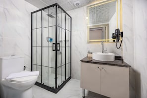 A modern bathroom with a glass-enclosed shower, a white toilet, a sink with a marble countertop, and a mirror with a light fixture.