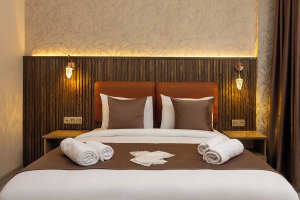 A neatly made bed with two pillows, a brown bedspread, and two rolled towels on the side.
