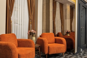 A hotel lobby with a checkered floor, a seating area with orange upholstered chairs, a vase with flowers, and a large mirror reflecting the room.