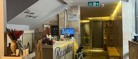 Un área de recepción con mostrador de mármol, una mesa de recepción, extintor, estatua y un cartel que indica la salida.
