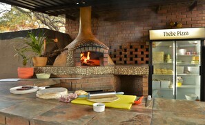 An outdoor pizza oven with a brick facade, a rolling pin, pizza dough, and a sign that reads "Thebe Pizza". - Thebe River Safaris (Kasane)