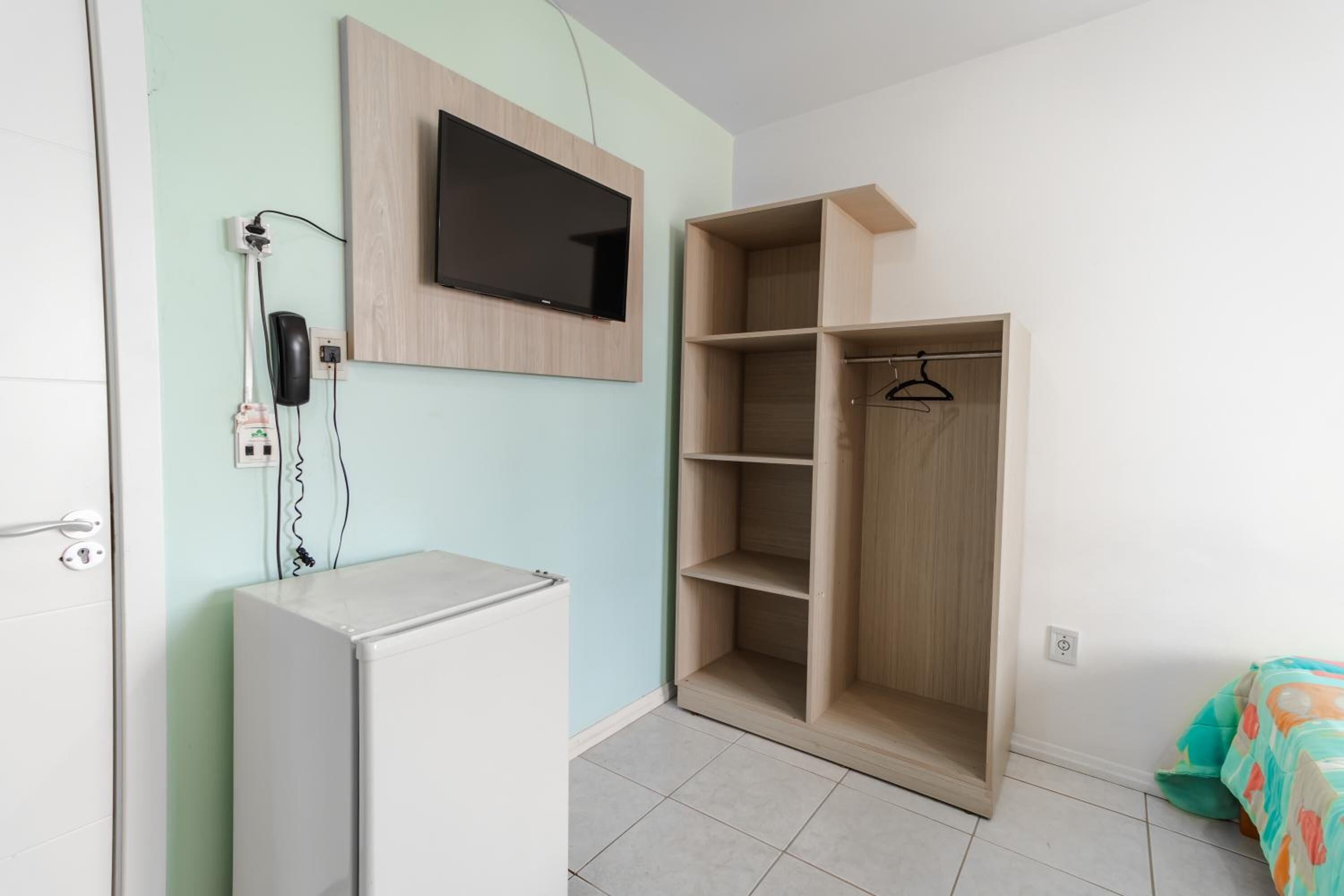 A room with a white refrigerator, a wooden wardrobe, a mounted TV, and a power outlet with a phone.
