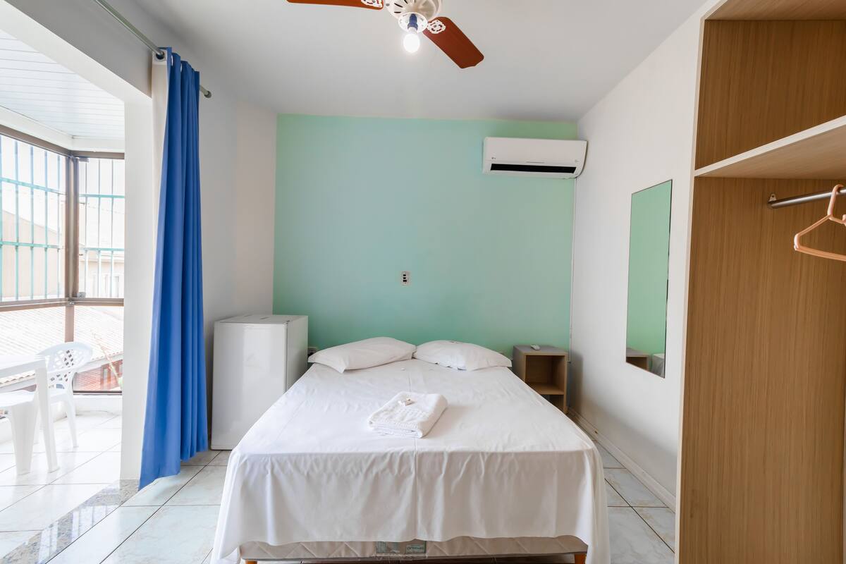 Une chambre avec un lit, un ventilateur de plafond, un appareil de climatisation, une fenêtre avec des rideaux bleus et un balcon avec une chaise.