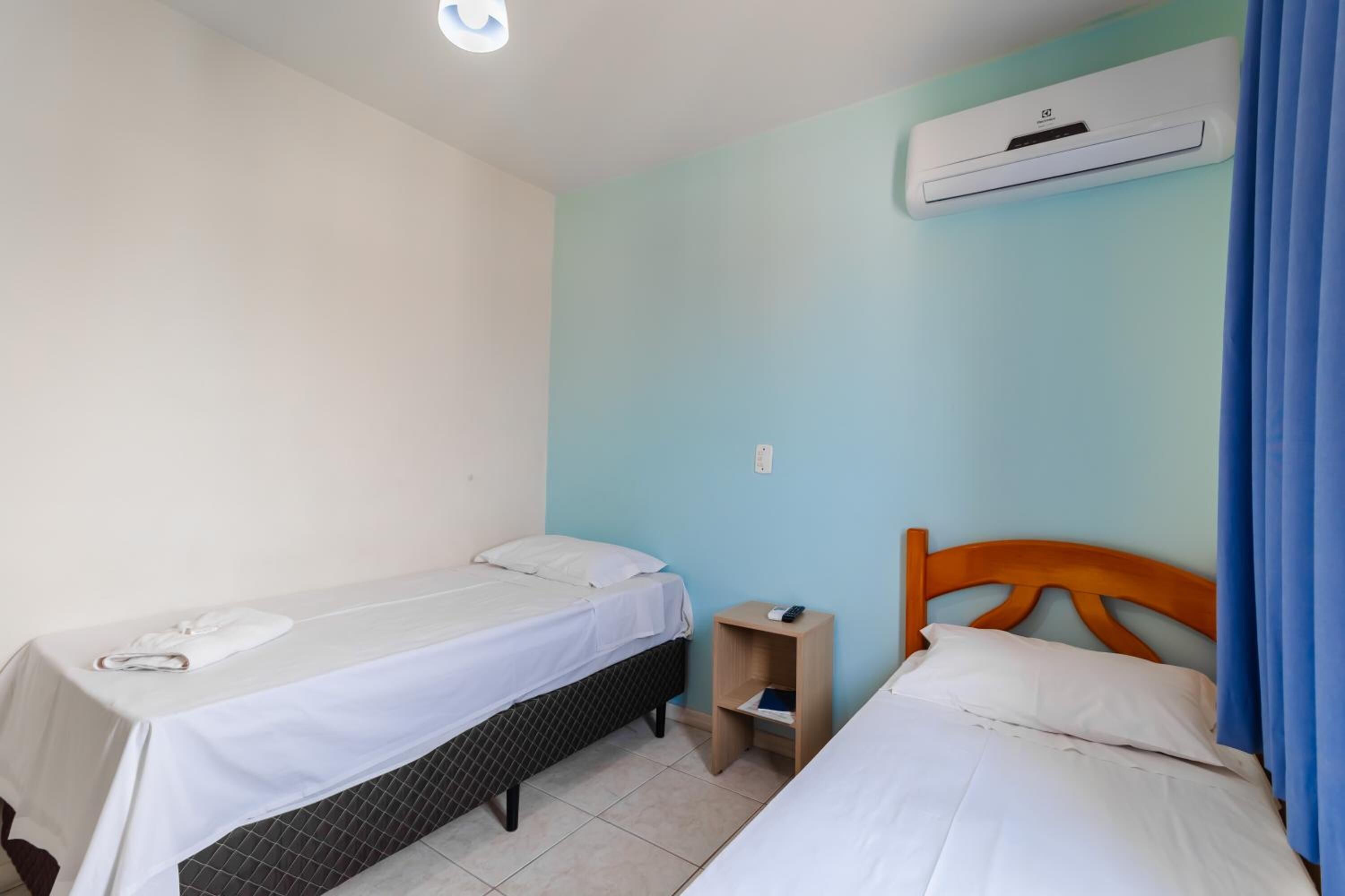 Quarto com duas camas, uma pequena mesa de cabeceira de madeira e um ar condicionado na parede.