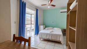 Une pièce comprenant un lit, un bureau et une chaise. Il y a une fenêtre avec des rideaux bleus, un ventilateur de plafond et un appareil de climatisation blanc.