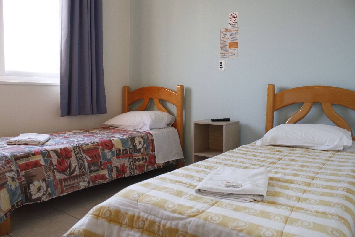 Une chambre avec deux lits simples, chacun doté d’une tête de lit en bois, d’une petite table de chevet et d’une prise de courant fixée au mur.