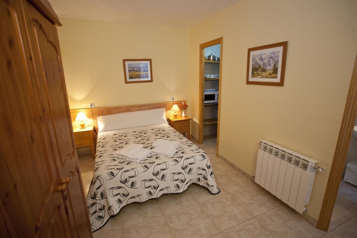 A bedroom with a bed, two bedside tables with lamps, a wooden wardrobe, a radiator, and two framed pictures on the wall.