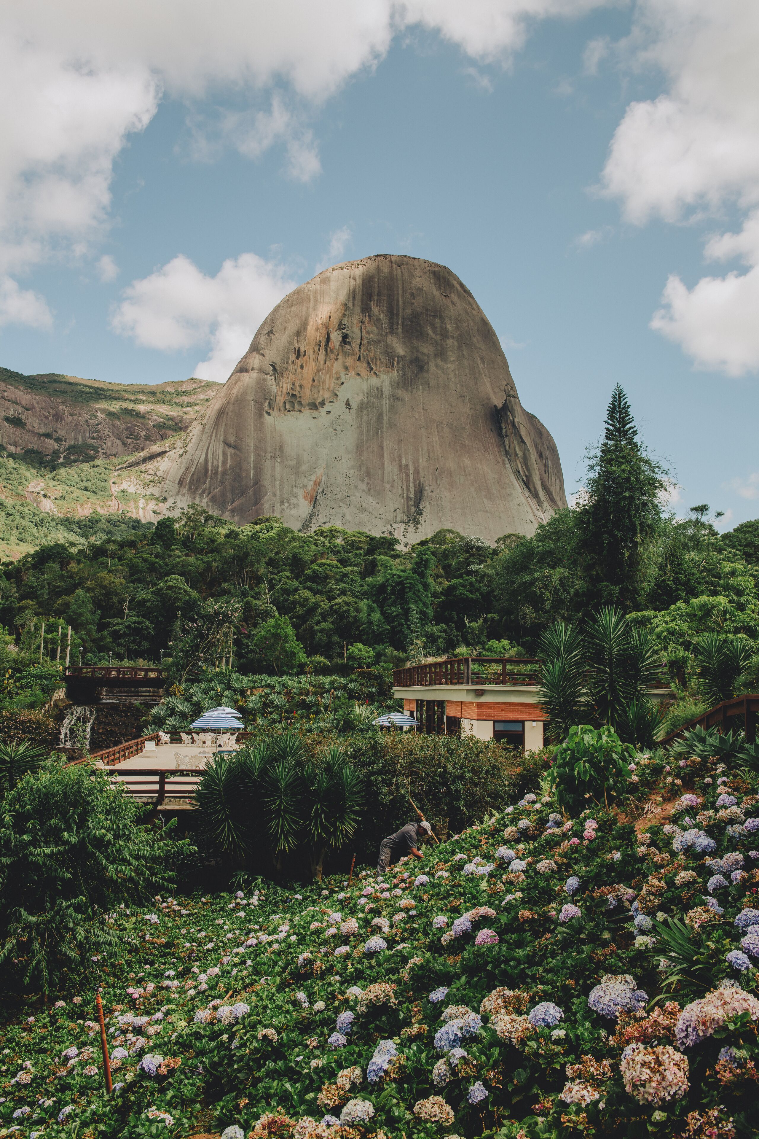 A natural landscape with a large rock formation, lush greenery, and a small building with a blue roof.
