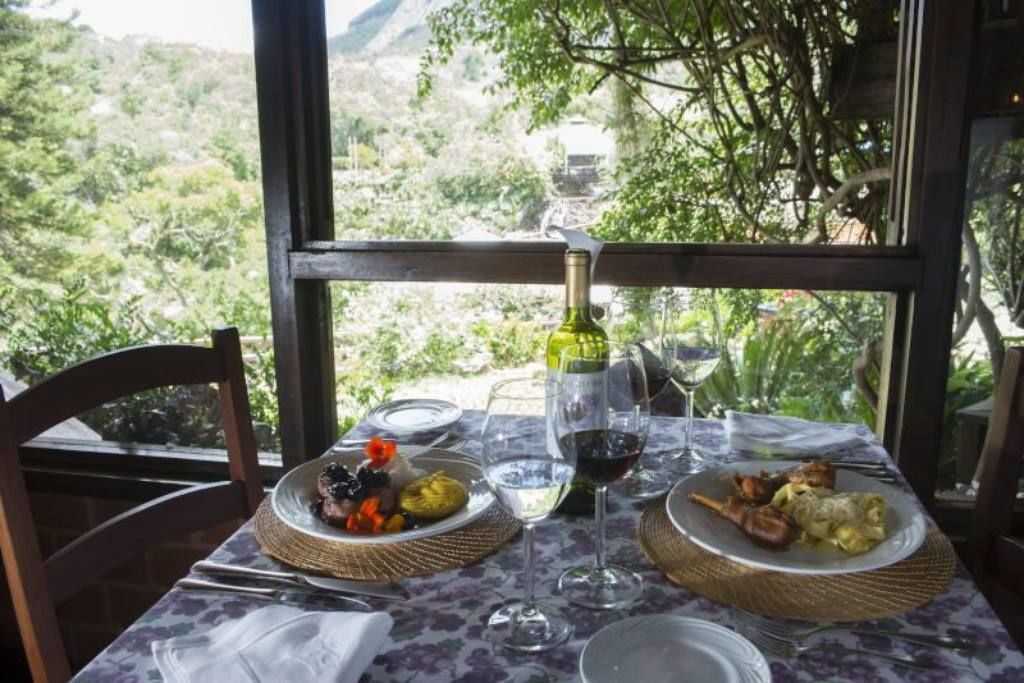 Ein Tisch reichhaltig mit Speisen, Weingläsern und einer Weinflasche gedeckt, mit Blick auf eine Berglandschaft.