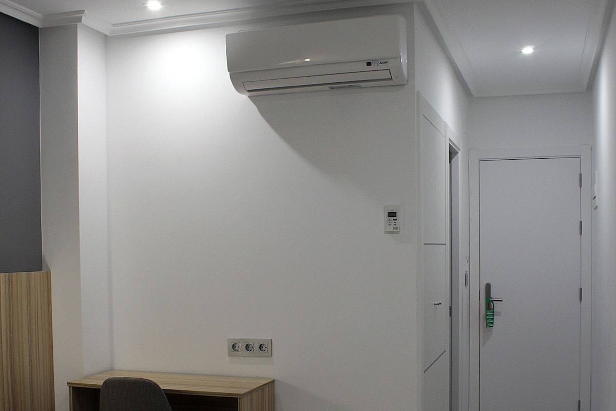 A hotel room with a bed, a desk, a chair, and a small table. There is an air conditioning unit on the wall and a door with a handle.
