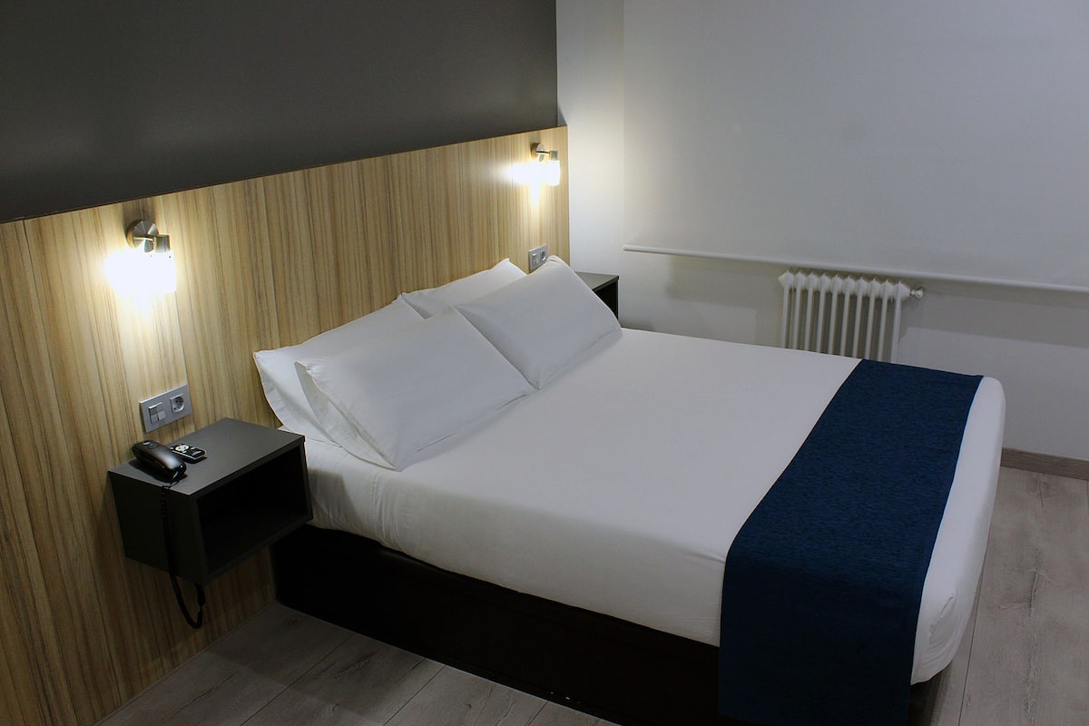 A neatly made bed with white linens and a blue blanket, a bedside table with a phone and a lamp, and a wall-mounted light fixture.