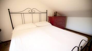 Una cama individual con estructura metálica y ropa blanca, un aparador rojo con cajones, y una pared blanca lisa.