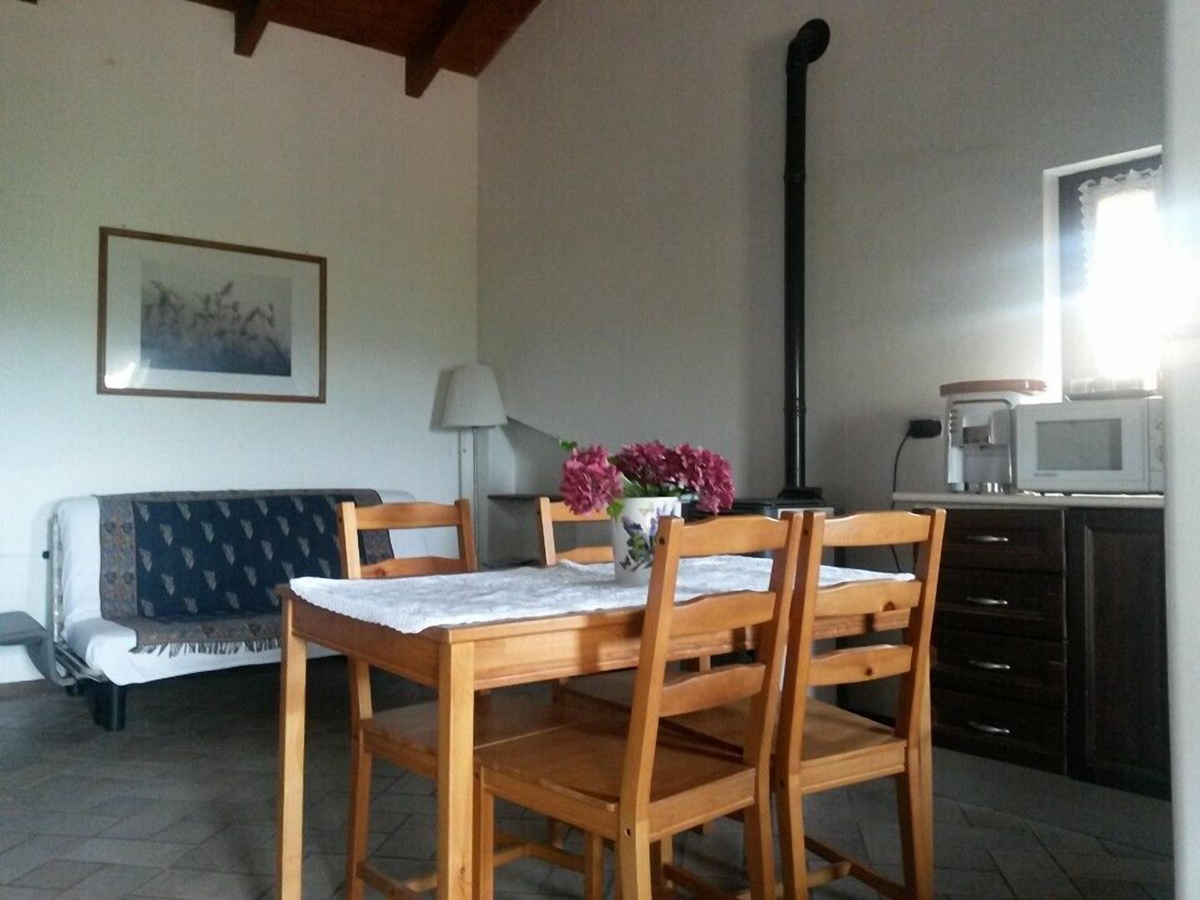 Ein Essbereich mit Holztisch und Stühlen, einer Couch, einer Lampe, einer Vase mit Blumen und einem Bild an der Wand. Zusätzlich ein Küchenbereich mit Mikrowelle und Herd.