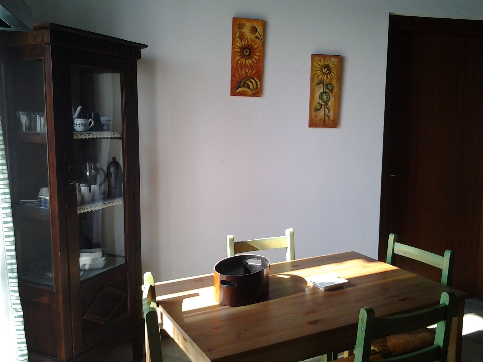 Un comedor con una mesa y sillas de madera, un armario con puertas de vidrio y dos cuadros en la pared.