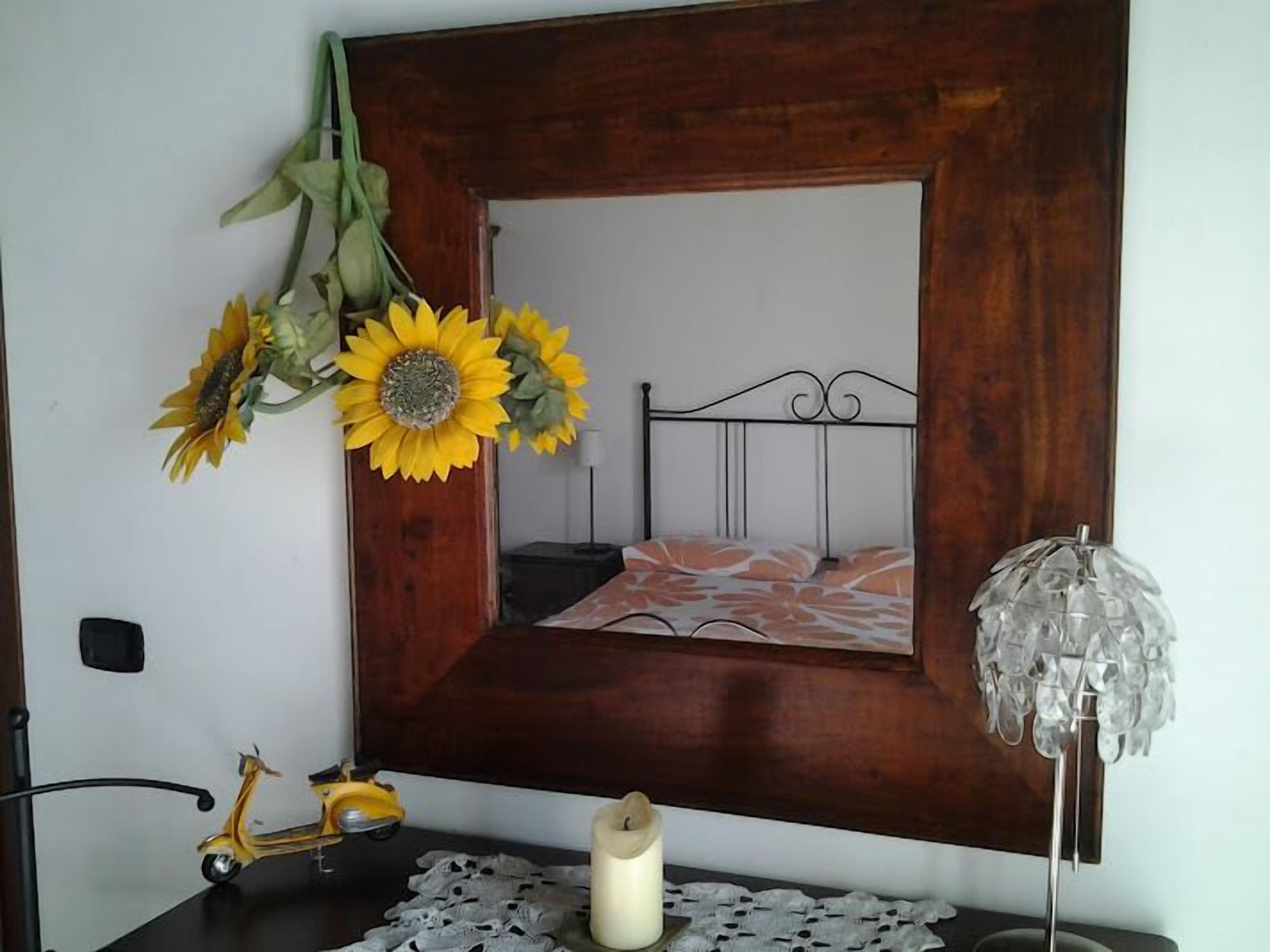 Un dormitorio con cabecera de madera, cama con ropa de cama floral, espejo con marco de madera, una mesita con un adorno y una lámpara de pared.