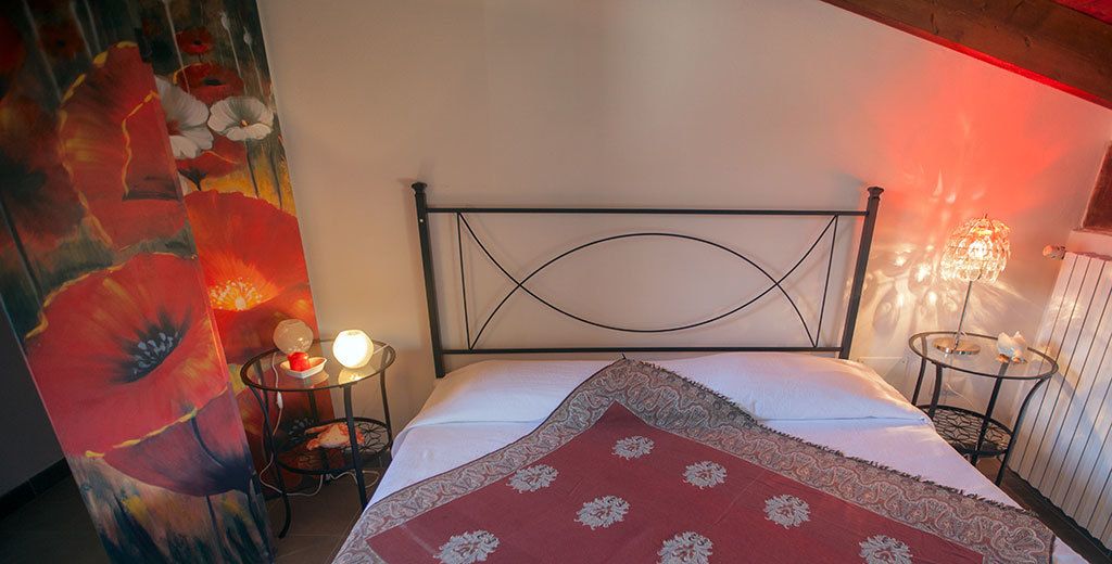 A bedroom with a metal headboard, a bed with a red and white patterned bedspread, two bedside tables with lamps, and a floral painting on the wall.