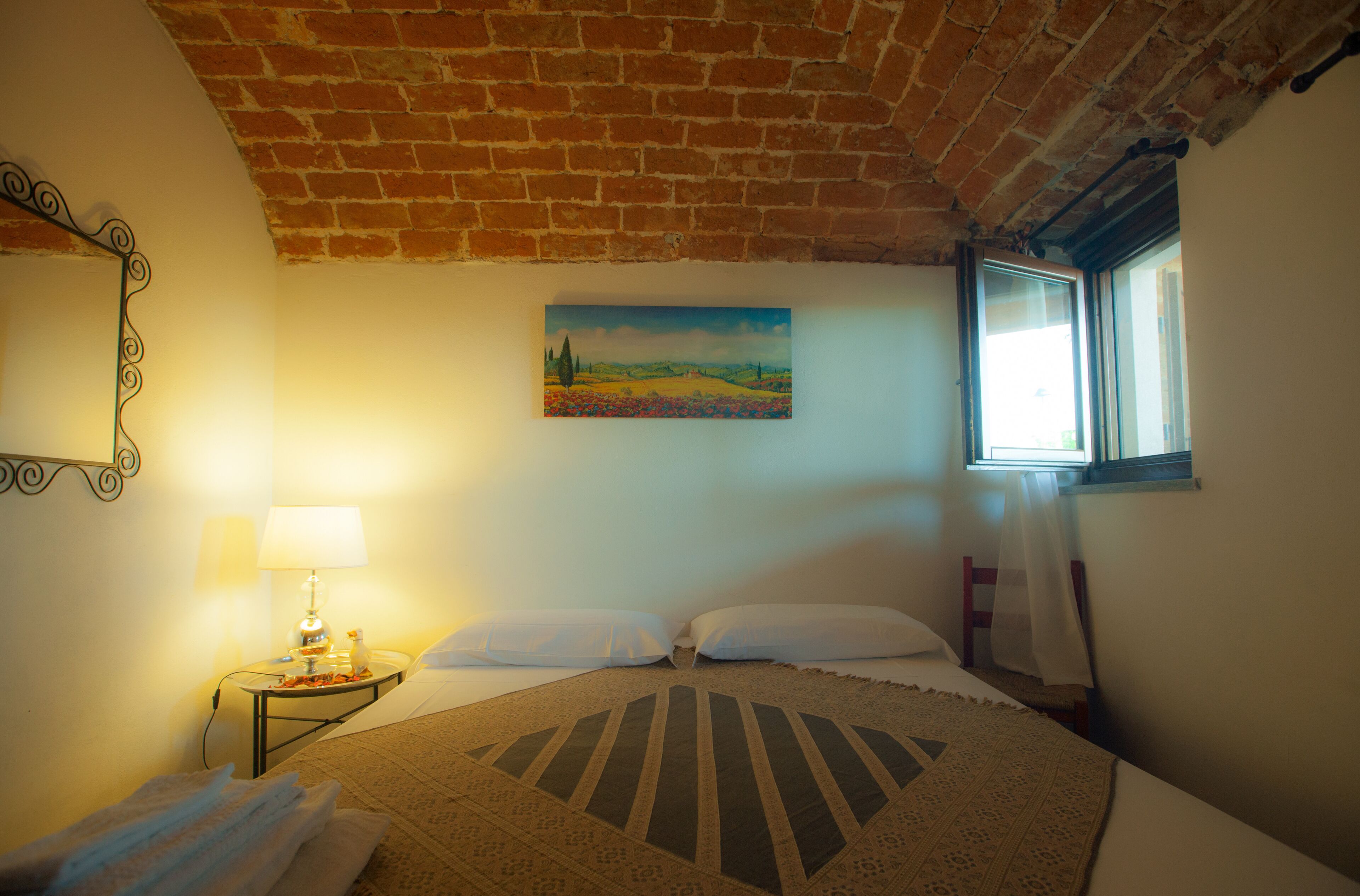 Un dormitorio con techo de ladrillo, un cuadro de un paisaje, una ventana con vistas, una cama con colcha estampada y una mesita de noche con lámpara y una botella.
