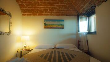 Un dormitorio con techo de ladrillo, un cuadro de un paisaje, una ventana con vistas, una cama con colcha estampada y una mesita de noche con lámpara y una botella.