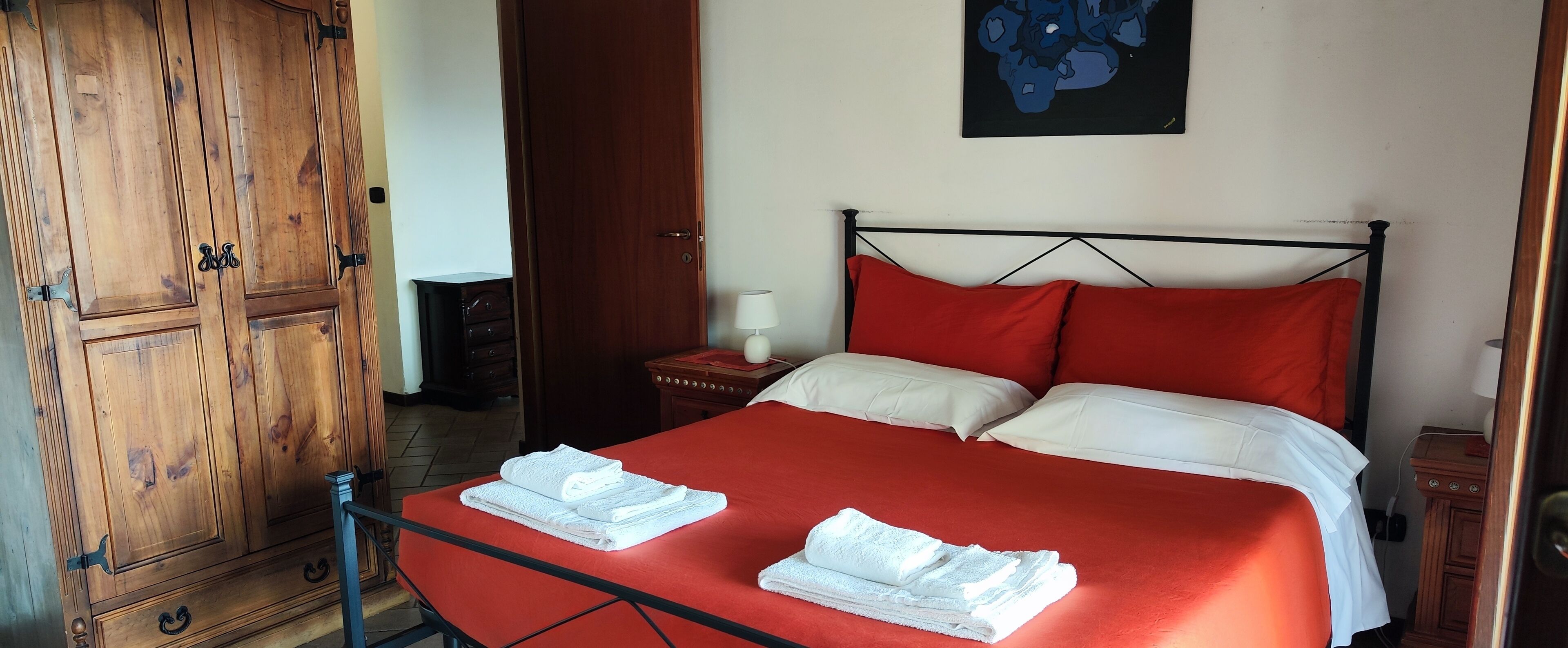 A neatly made bed with red bedding and white pillows, a wooden wardrobe, and a small bedside table with a lamp.
