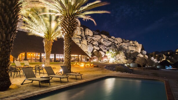 A nighttime pool area with lounge chairs, a palm tree, and a building with a stone facade.