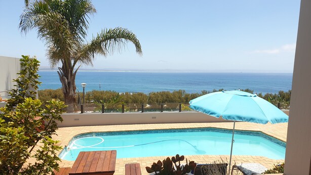A pool area with a view of the ocean, wooden furniture, and a potted plant. - 59LongSantos (Mossel Bay)