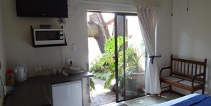 A room with a bed, a bench, a microwave, and a sliding glass door leading to a garden. - Angelica Guest House (Boksburg)