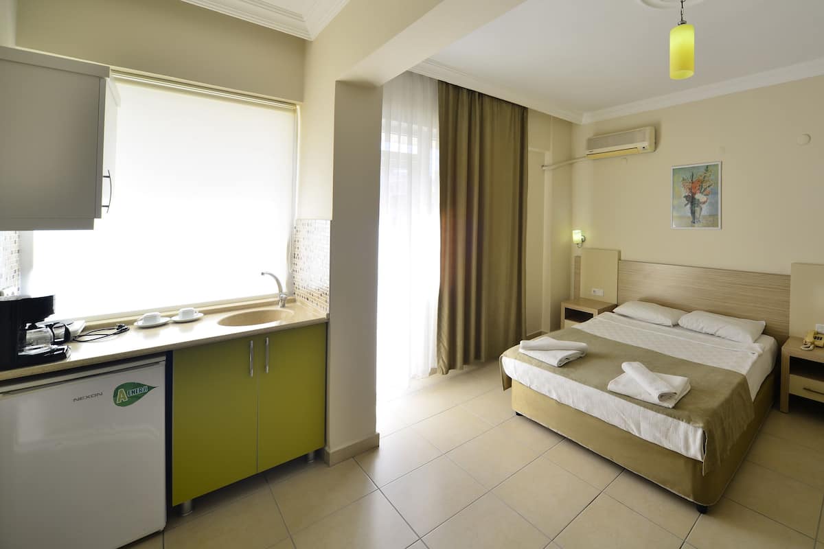 Habitación de hotel con cama, mesitas de noche, escritorio, espejo, ventana con cortinas, lavamanos y kitchenette.