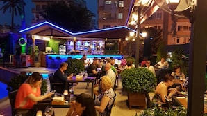 An outdoor dining area with tables and chairs, a bar in the background, and surrounding greenery. - Prince Apart Hotel (Alanya)