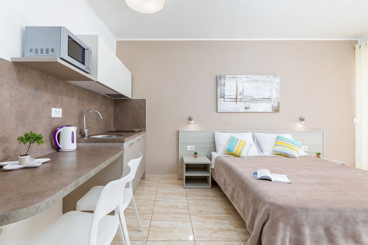 A modern bedroom with a bed, bedside tables, a nightstand, a small desk, a chair, a microwave, a kettle, and a potted plant.