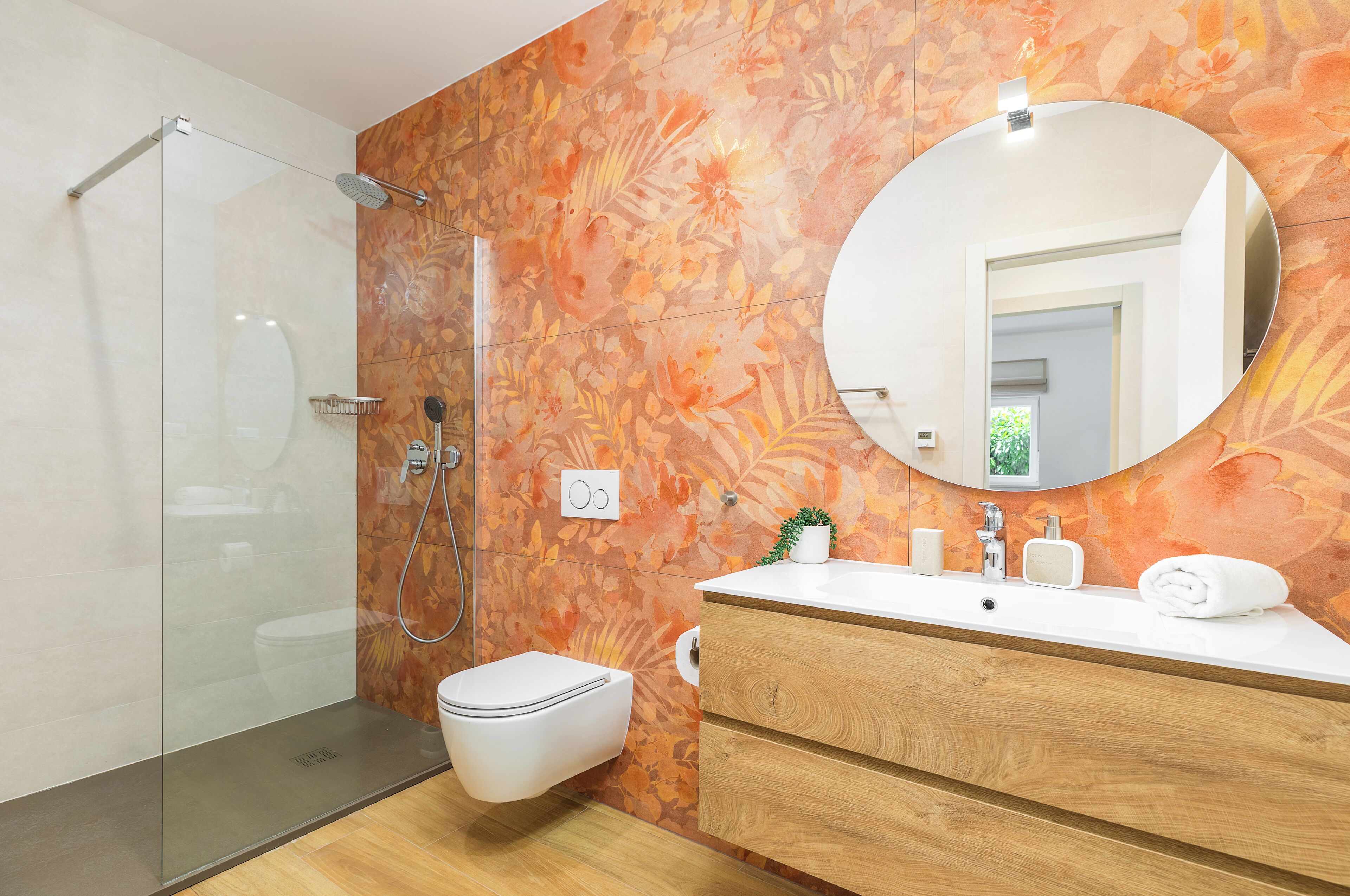 A modern bathroom with a glass shower enclosure, a toilet, a wooden vanity with a mirror, and a floral-patterned wall.