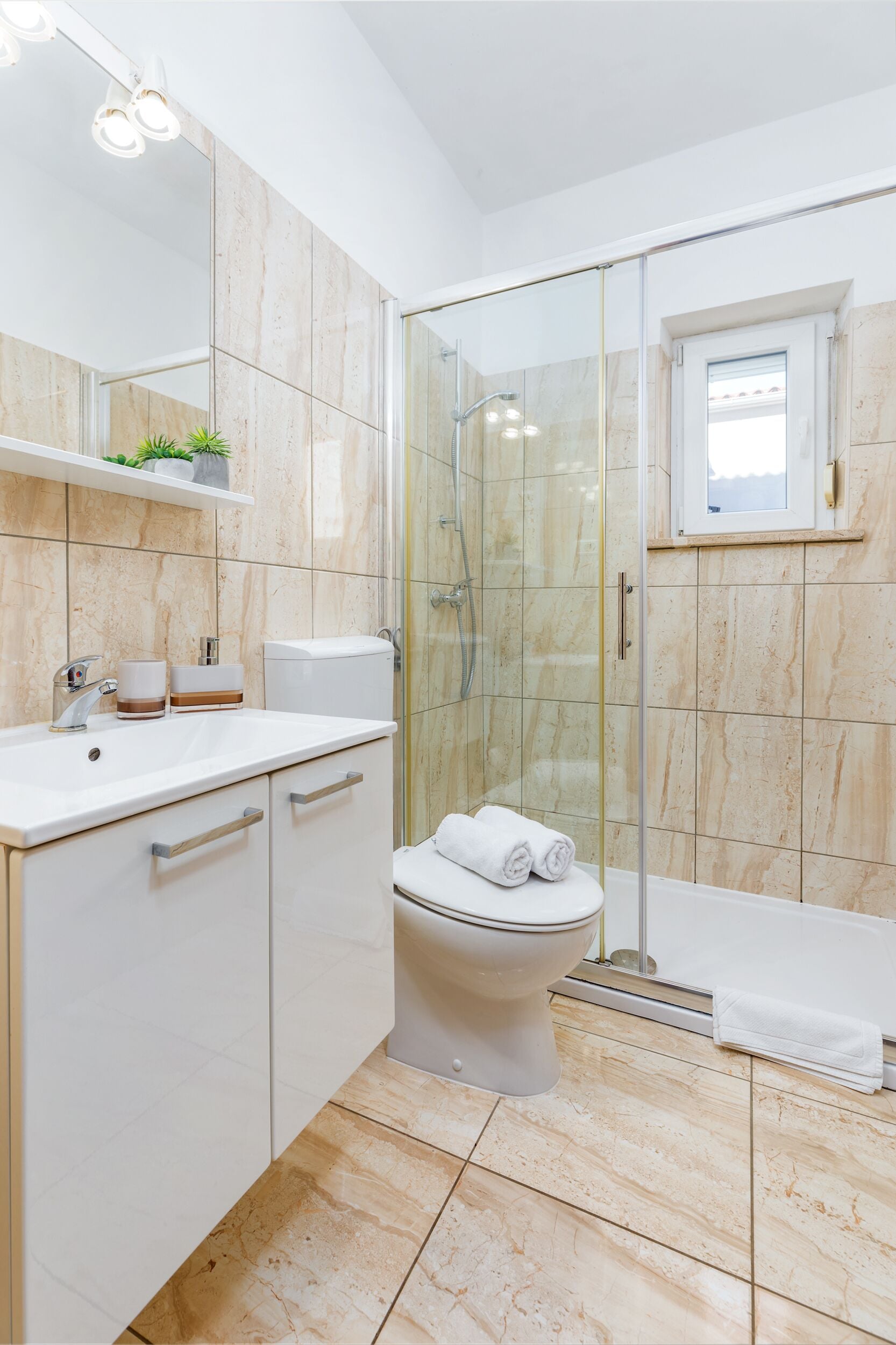 A modern bathroom with a glass-enclosed shower, a toilet, a sink with a mirror, and a window above the toilet.
