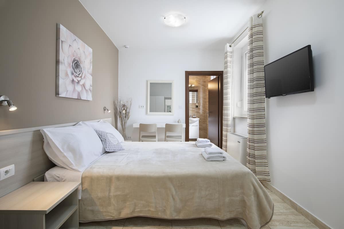 A hotel room with a bed, bedside table, a flat-screen TV mounted on the wall, a bathroom with a mirror, and a shower area.