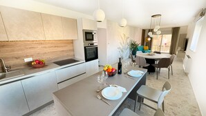 A modern kitchen with a central island, built-in oven, and microwave. There is a dining area with a round table and chairs, and a living space with a sofa and TV.