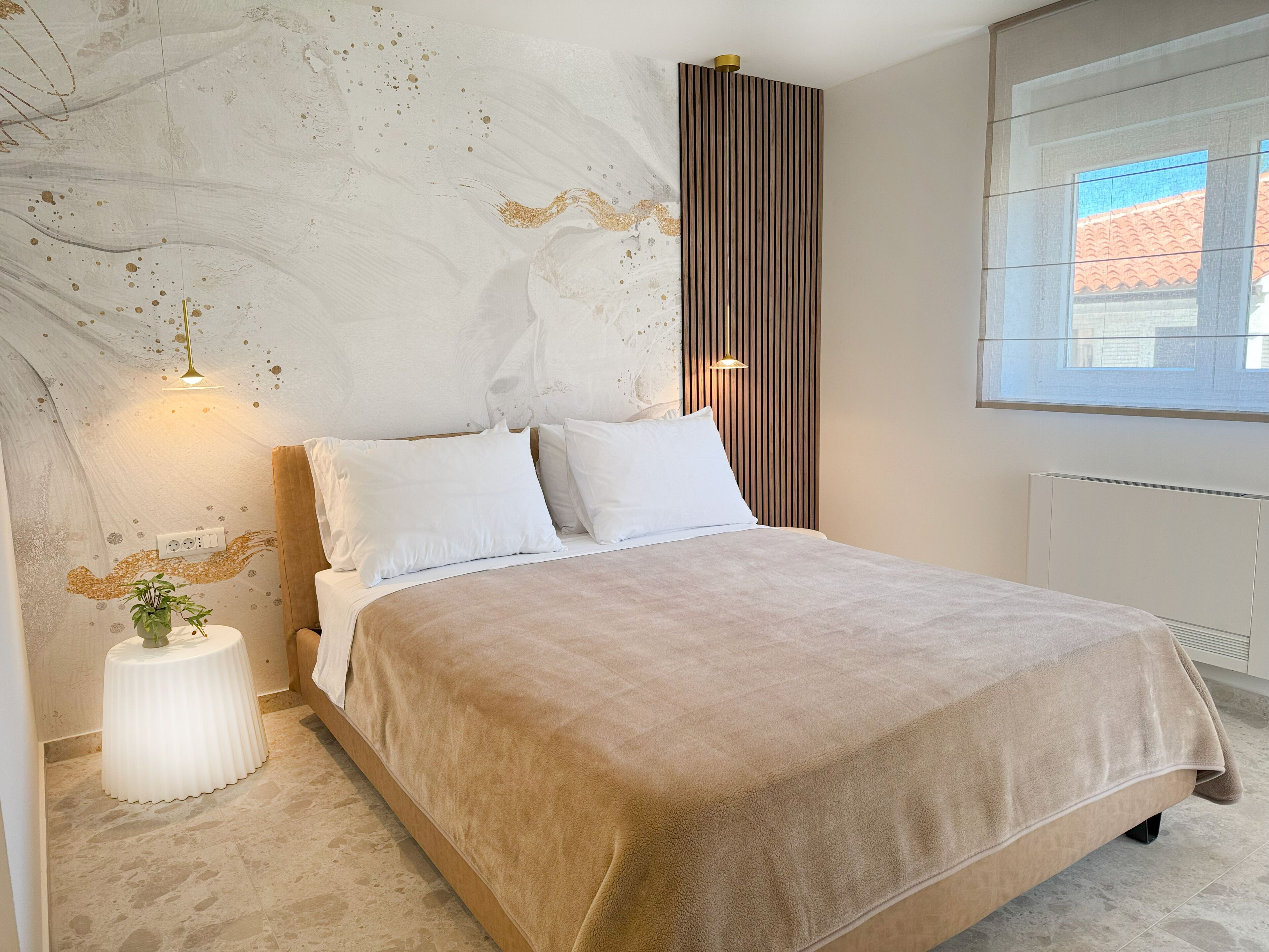 A bedroom with a bed, white pillows, a beige blanket, a small white side table with a plant, and a window with a view of buildings.