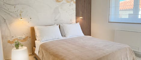 A bedroom with a bed, white pillows, a beige blanket, a small white side table with a plant, and a window with a view of buildings.