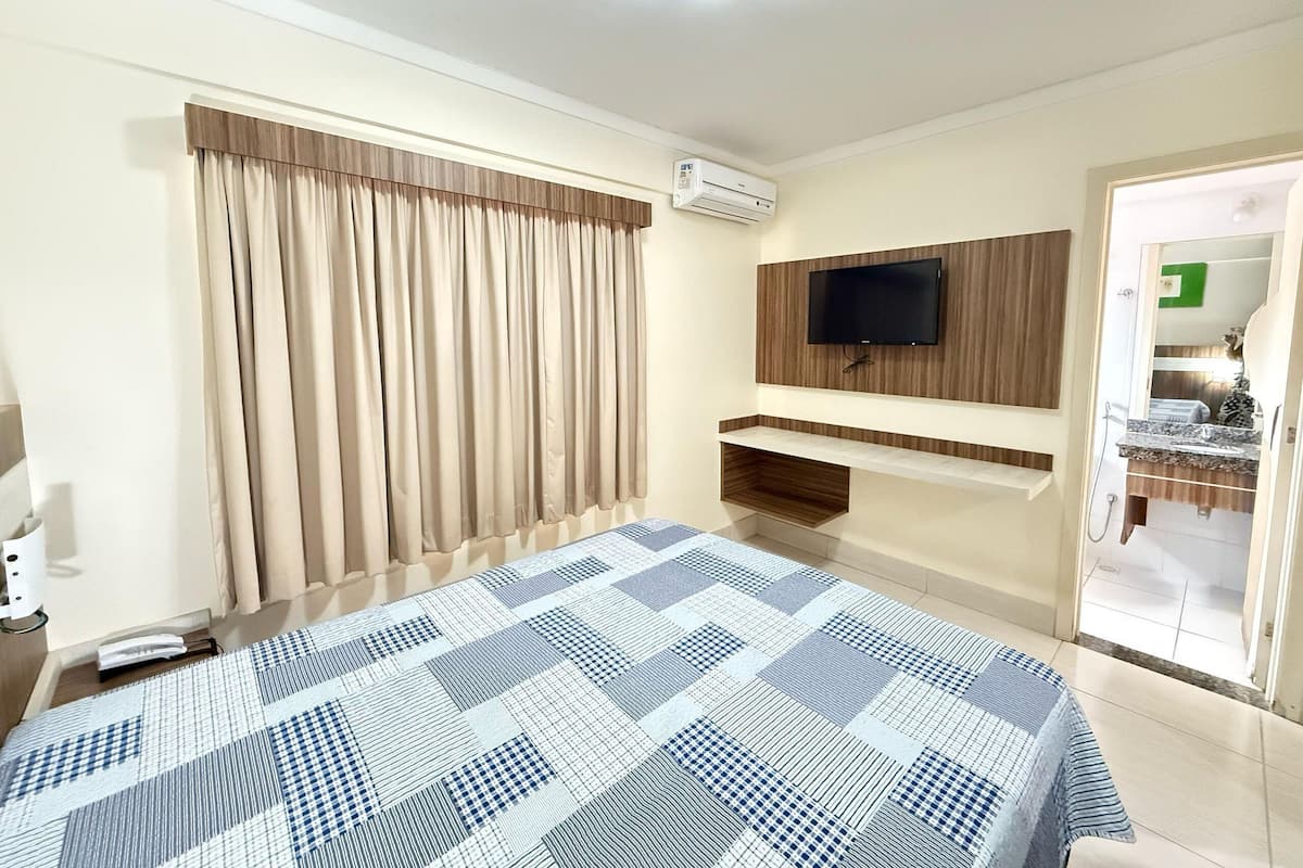 A hotel room with a bed, a TV mounted on the wall, an air conditioning unit, and a bathroom visible through an open door.