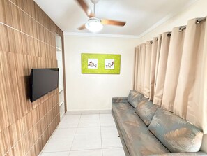 A room with a flat-screen TV mounted on a wooden wall, a ceiling fan, a sofa with a leaf pattern, and two framed pictures on the wall. - Lacqua DiRoma IV Via Caldas (Caldas Novas)