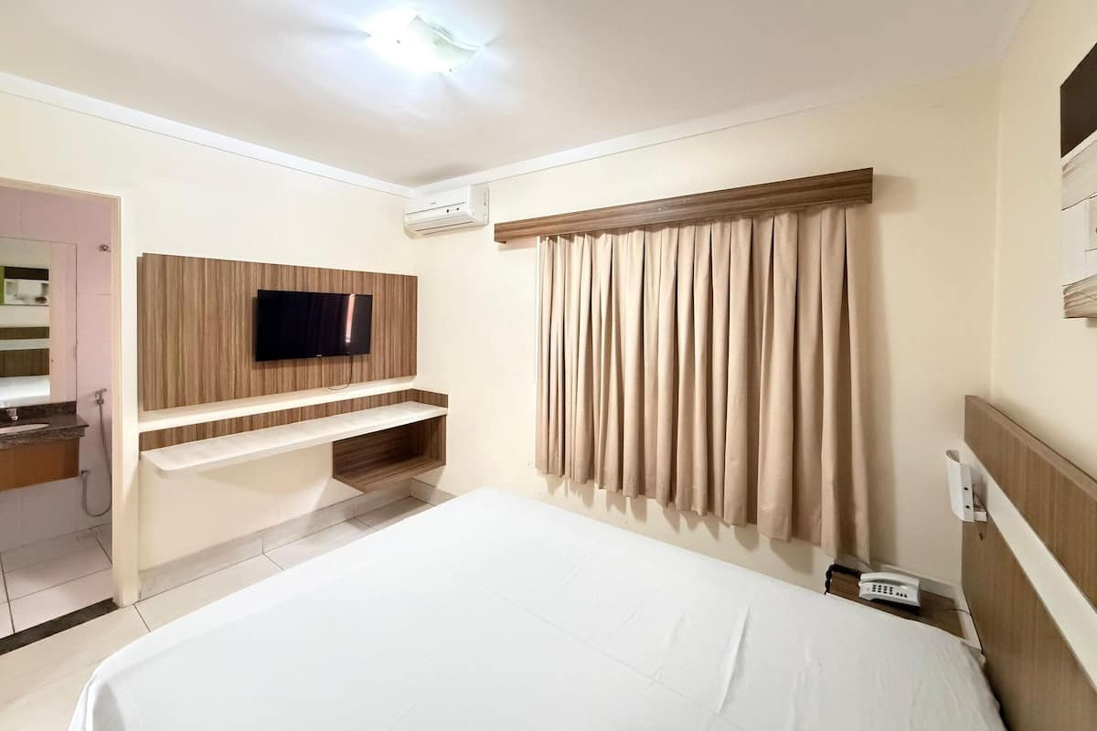 A hotel room with a bed, a TV mounted on a wooden panel, a bathroom with a sink and mirror, and a curtain on the window.