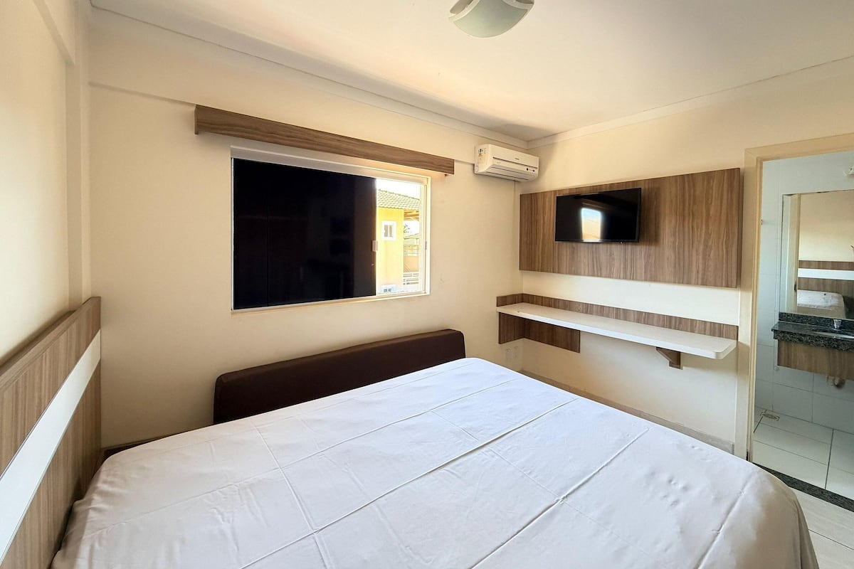 A hotel room with a bed, a TV mounted on the wall, a window, and an air conditioning unit.