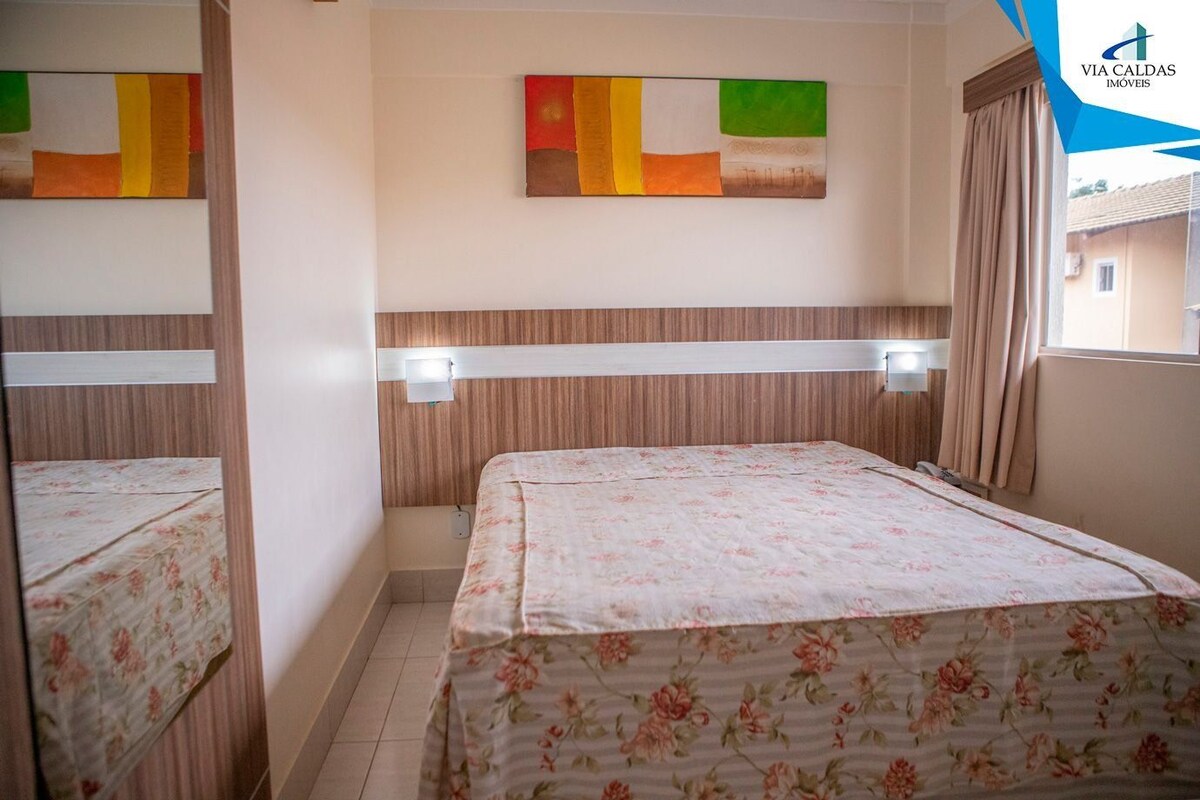 A hotel room with a bed, a wall-mounted artwork, a window with curtains, and a visible logo.