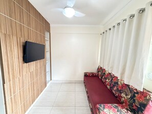 Une pièce avec un mur en bois, un téléviseur à écran plat, un ventilateur de plafond, un canapé recouvert d’un tissu à motifs floraux et une fenêtre avec des rideaux.