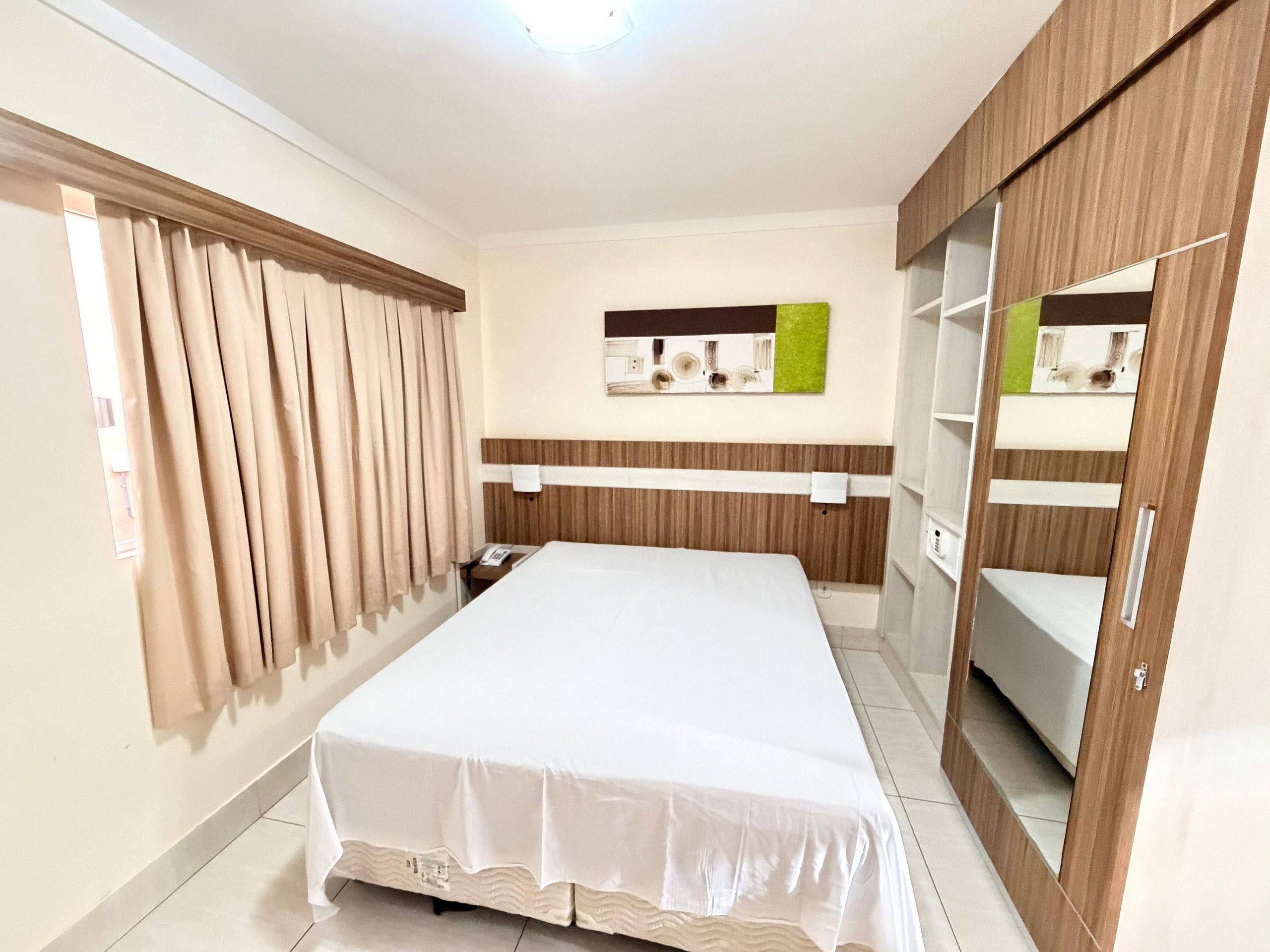 A hotel room with a bed, curtains, a bedside table, a mirror, and a wardrobe.