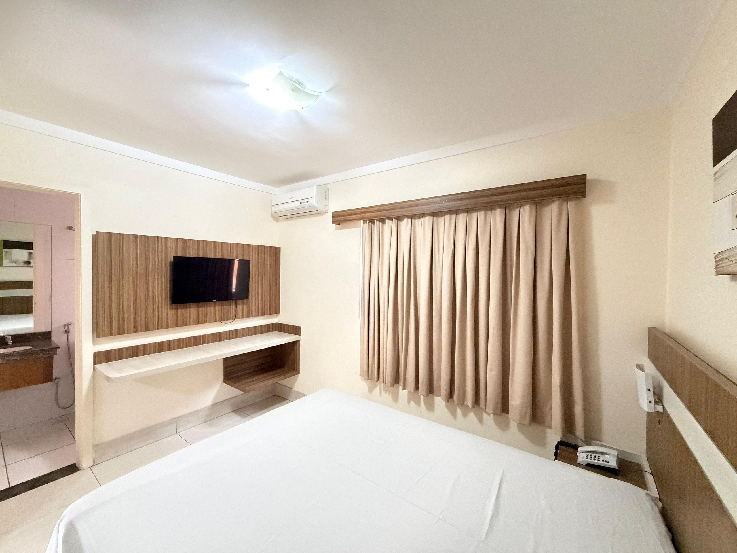 Habitación de hotel con cama, televisión instalada en un panel de madera, baño con lavamanos y espejo, y cortina en la ventana.