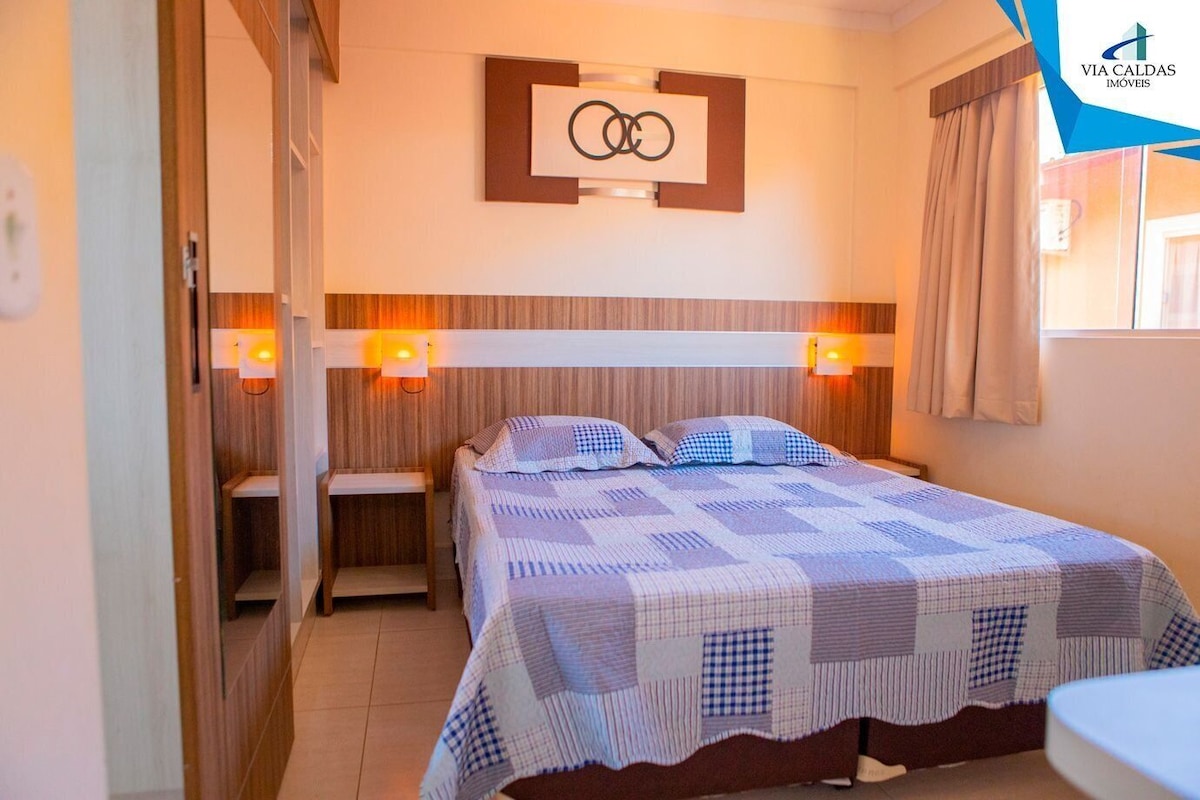 Un dormitorio con cama, dos mesitas de noche, una lámpara de pared y una ventana con cortinas.