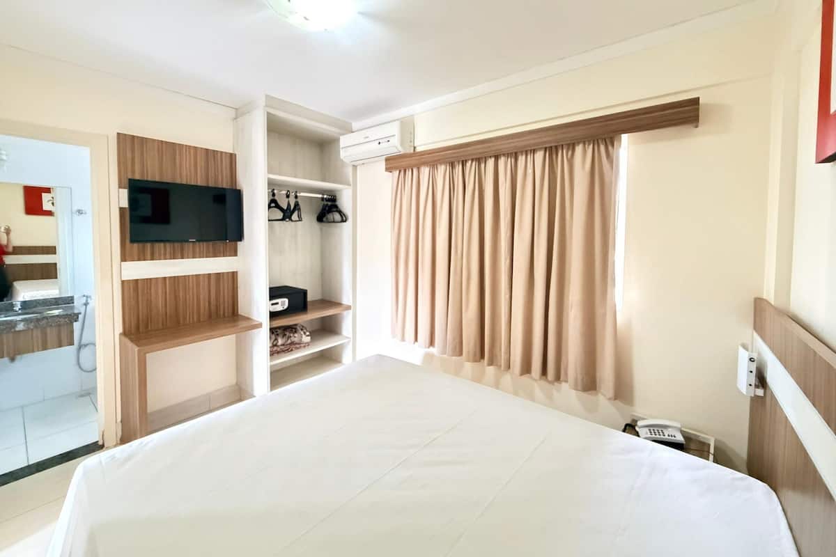 A hotel room with a bed, a TV mounted on a wooden panel, a wardrobe with hanging shoes, a bathroom with a mirror, and a telephone.